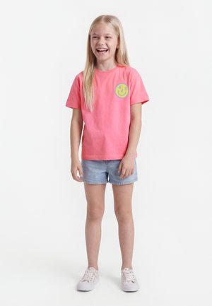 Niña sonriente de cabello largo y rubio, vestida con una camiseta rosa brillante con gráfico de sonrisa, pantalones cortos de mezclilla claros y zapatillas blancas.