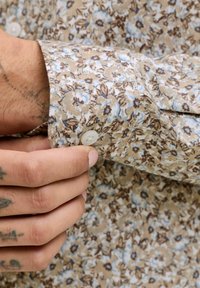 Main tatouée en train de boutonner le poignet d'une chemise à manches longues beige à motif floral bleu.