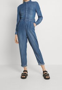 Mono de denim en azul medio con mangas largas, cuello, bolsillos en el pecho, cintura elástica y piernas ajustadas con dobladillos en los puños. Sandalias negras.
