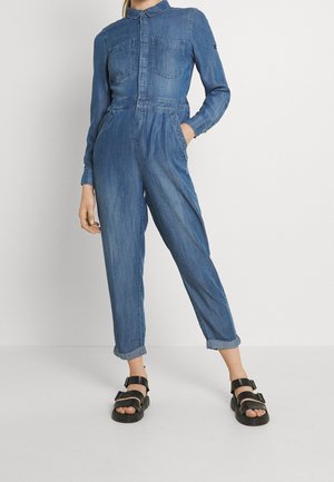 Mono de denim en azul medio con mangas largas, cuello, bolsillos en el pecho, cintura elástica y piernas ajustadas con dobladillos en los puños. Sandalias negras.