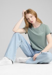 Svetlozelené bavlnené tričko s vyšitým logom "Puma", skombinované s voľnými svetlomodrými džínami a bielymi teniskami, sediaca póza na neutrálnom pozadí.