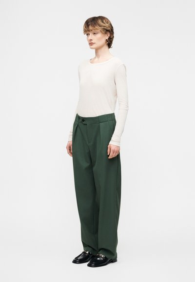 Γυναίκα με κοντά μαλλιά που φοράει λευκό πουκάμισο με μακριά μανίκια, σκούρο πράσινο παντελόνι με φαρδιά πατζάκια, μαύρα loafers και χρυσούς κρίκους σκουλαρίκια, στέκεται πλαγίως.