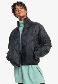 Black puffer jacket with a quilted pattern and high collar, featuring a front zipper. Worn over a light green sweater.