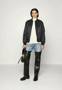 Casaco bomber preto sobre um suéter branco de gola alta, combinado com jeans azuis e calças pretas de imitação de couro, finalizado com botas pretas robustas e uma pequena bolsa.