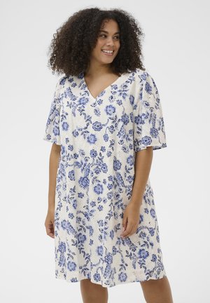 Woman with curly hair wearing a white knee-length dress with blue floral patterns, smiling and looking to the side.