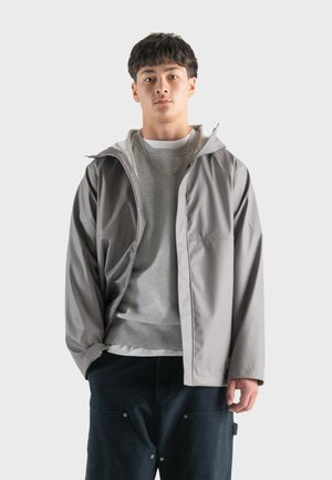 Jeune homme portant une veste à capuche gris clair sur un sweat gris et un pantalon noir, se tenant devant un fond blanc uni.