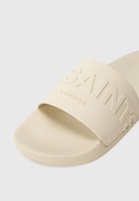 Béžová šľapka s textúrovanou stielkou a širokým remienkom s vyrazeným textom „SAINTS LONDON“ na hornej strane.