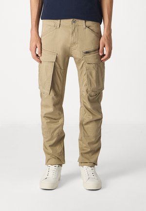 Pantalon cargo beige avec plusieurs poches, y compris des poches à rabat et zippées, porté avec des baskets blanches et un haut foncé.