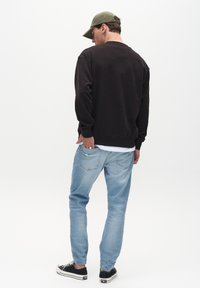 Sudadera negra con cuello redondo, jeans azules desgastados y zapatillas negras. Lleva una gorra verde oliva, visto desde atrás.