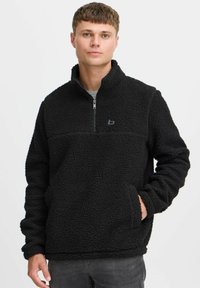 Pull en polaire noir avec un col zippé mi-hauteur, un col haut et une poche, présentant une surface texturée et un logo discret sur la poitrine.