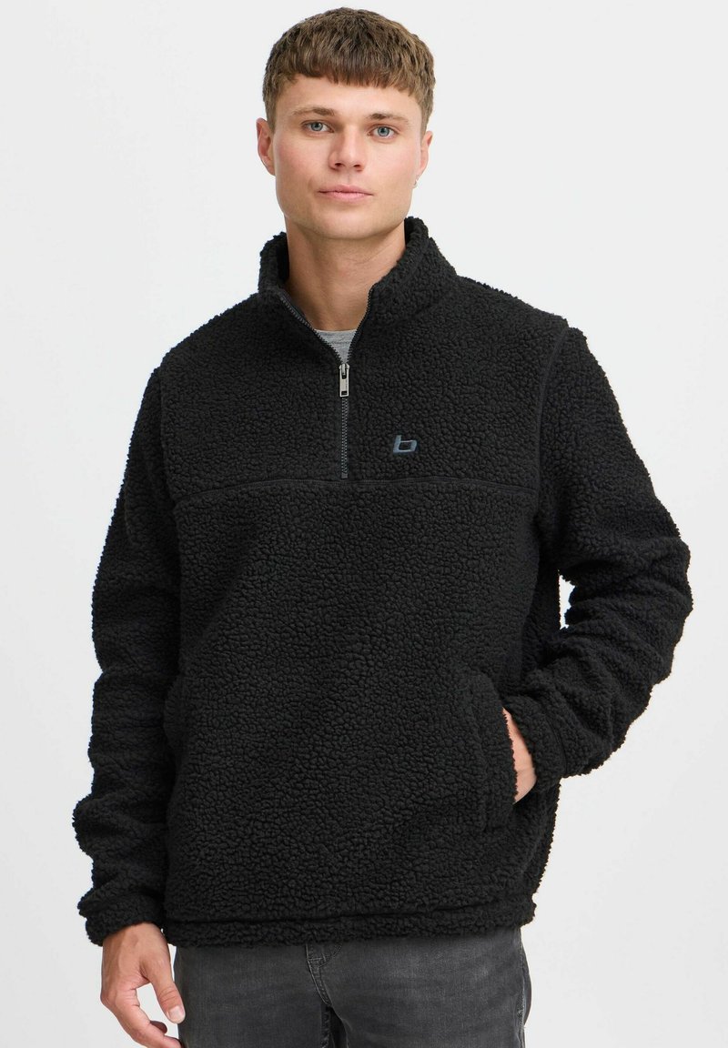 Pull en polaire noir avec un col zippé mi-hauteur, un col haut et une poche, présentant une surface texturée et un logo discret sur la poitrine.