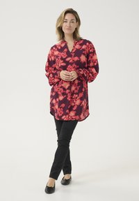 Czerwona bluzka z długim rękawem w kwiatowy wzór z lekkiego materiału, zestawiona z czarnymi slim-fit spodniami i czarnymi płaskimi butami na neutralnym tle.