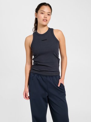 Kobieta z ciemnymi włosami związanymi w kucyk, ubrana w czarny bezrękawnik prążkowany i granatowe spodnie, stojąca z jedną ręką w kieszeni.