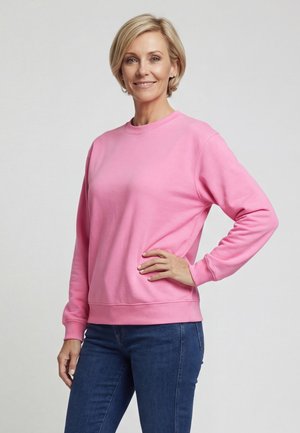 Mosolygó nő rövid szőke hajjal, rózsaszín pulóvert és kék farmert viselve, egy kézzel a csípőjén áll, egyszerű háttér előtt.