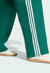 Zelené sportovní kalhoty s bílými bočními pruhy, spárované s bílými teniskami s béžovou podrážkou a texturovaným detailem.