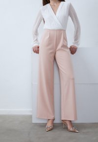 Blusa blanca texturada con lunares y un escote en V profundo, combinada con pantalones de pierna ancha en color rosa y tacones transparentes decorados con lazos de joyas.