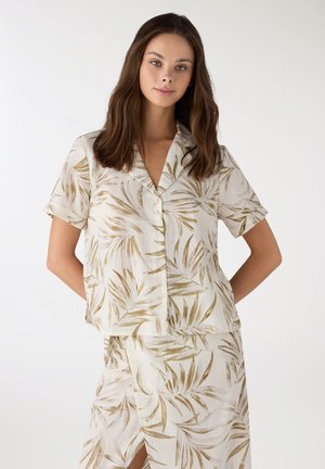 Giovane donna che indossa una camicia crema a maniche corte con bottoni e una gonna coordinata con motivi di foglie marrone chiaro, posando davanti a uno sfondo semplice.