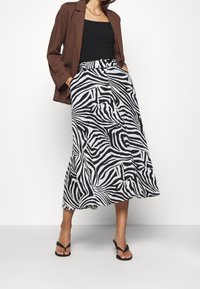 Zwart-witte zebra print midi-rok met een uitlopende vorm, voorzien van een tailleband en knoopdetail. Gecombineerd met een bruine blazer en een zwarte top.