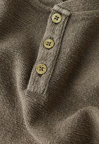 Tissu marron texturé avec trois boutons en bois clair alignés verticalement sur une patte de vêtement en tricot.