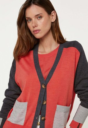 Vrouw met lang bruin haar, gekleed in een rode en donkergrijze cardigan met knopen, met lichtgrijze zakken en manchetten.