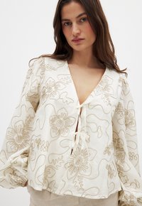 Embroidered cream blouse with a deep V-neck, tied closure, and puffy sleeves. Features floral patterns in beige thread. Textured fabric.
