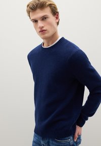 Marineblauer, strukturierter Strickpullover mit Rundhalsausschnitt und gerippten Bündchen, darüber ein weißes T-Shirt mit Rundhalsausschnitt, kombiniert mit blauen Jeans.