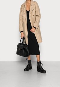Beiger Trenchcoat über einem schwarzen Kleid, kombiniert mit schwarzen Schnürstiefeln und einer strukturierten schwarzen Handtasche mit Obergriff.
