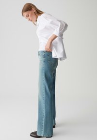 Jeans chiari con design a gamba dritta, abbinati a una camicia bianca con bottoni. Caratteristiche includono una vita media e tasche posteriori.