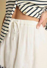Biele ľanové nohavice s elastickým pásom na šnúrku, ktoré majú voľný strih a jemnú textúru. Čiastočne je viditeľný pruhovaný top v námorníckej modrej a krémovej farbe.
