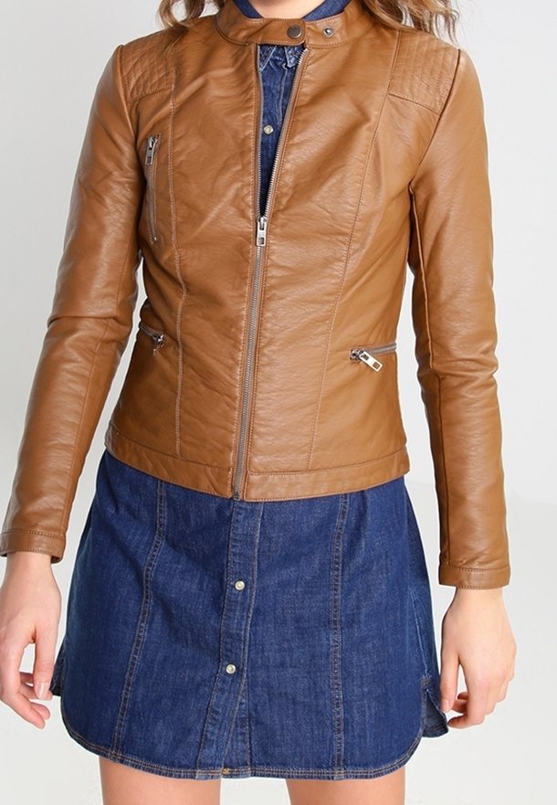 Femme portant une veste en cuir marron sur une jupe en denim bleu boutonnée, se tenant contre un fond clair.