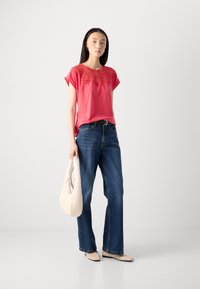 Chemise rose avec des broderies rouges complexes, associée à un jean bleu à jambes larges. Le mannequin tient un sac couleur crème et porte des ballerines de couleur neutre.