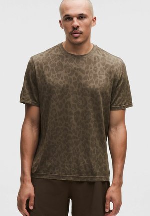 Uomo con indosso una camicia verde oliva a maniche corte con un sottile motivo a leopardato, in piedi davanti a uno sfondo chiaro e semplice.