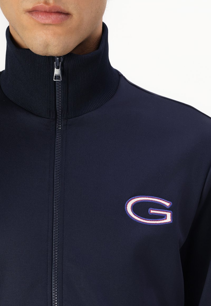 Chaqueta navy con cremallera y cuello alto, que presenta una cremallera frontal y un logo bordado con contorno blanco en el lado izquierdo del pecho. Textura suave.