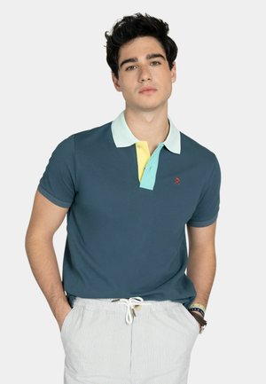 Giovane uomo con capelli scuri che indossa una polo blu con colletto color menta e giallo, e pantaloncini bianchi a righe con coulisse, mani in tasca.
