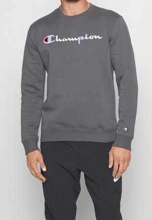 Hombre con sudadera gris de Champion con el logo en el pecho y la manga, combinada con pantalones negros de Nike que muestran el logo Swoosh cerca del bolsillo.