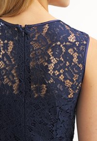 Navy lace dress with floral patterns, sheer texture, and solid neckline. Features a discreet zipper at the back, showcasing intricate detailing.