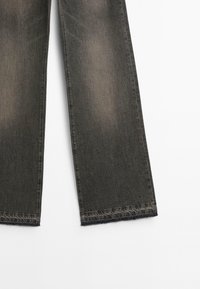 Dunkelgraue Jeans mit geradem Bein, versehen mit ausgewaschenen Stellen und rohen Kanten. Die Textur wirkt leicht abgetragen.