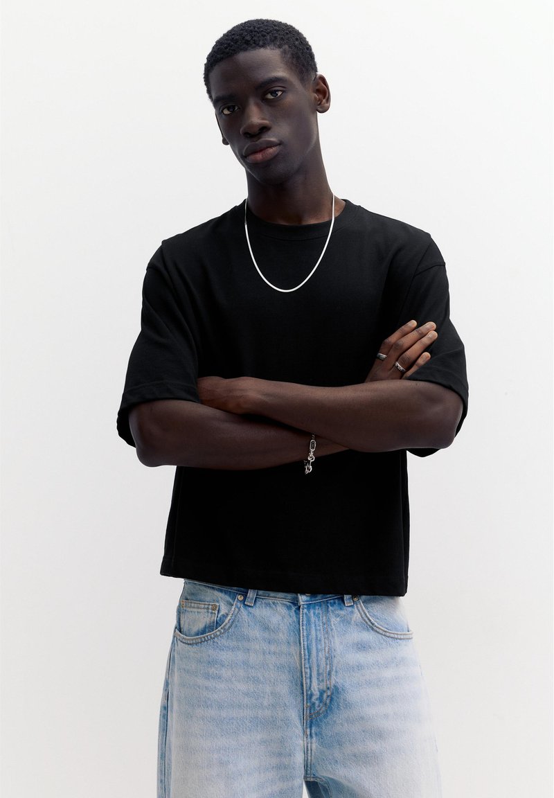 Jeune homme à la peau foncée portant un t-shirt noir, un jean bleu clair, un collier en argent, des bagues et un bracelet, debout les bras croisés sur fond blanc.
