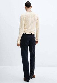 Kremowy dziergany sweter z liściastym wzorem, koronkowym wykończeniem na dole, w zestawie z ciemnoniebieskimi dżinsami. Całość dopełniają czarne buty na obcasie.