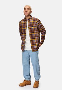 Ruutu- ja keltaisenruskea napillinen paita, valkoinen t-paita alla, vaaleansiniset farkut ja vaaleat saappaat. Sisältää rintataskun ja rentoa istuvuutta.