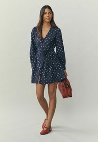 Marineblaues gepunktetes Kleid mit Kragen, langen Ärmeln und tailliertem Gürtel. Das Modell hält eine rote Handtasche und trägt rote Schuhe mit abgerundetem Zehenbereich.