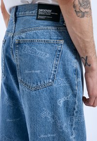 Los pantalones cortos de mezclilla azul claro presentan un acabado texturizado con un patrón de logotipos repetido. La cinturilla tiene una etiqueta negra de marca y detalles de costura.