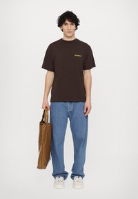 Braunes Baumwoll-T-Shirt mit "No Problemo" in Gelb bedruckt, kombiniert mit blauen Jeans und einer braunen Tragetasche. Weiße Sneakers runden den Look ab.