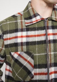 Casaco xadrez de lã em tons de verde, vermelho, preto e branco. Possui fecho de correr, colarinho espalhado e um bolso no peito com aba arredondada.