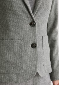 Veste grise à fines rayures avec un tissu texturé, deux boutons noirs et des poches latérales. Coupe cintrée avec un col à revers cranté.