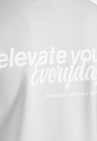 T-shirt bianca in cotone con testo grande e a contrasto che recita "eleva te stesso ogni giorno" con il nome del brand Smilodox sottostante.