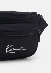 Mustavärinen kangas vyölaukku, jossa on useita vetoketjullisia taskuja ja valkoinen nimikkologo. Muotoilu on kompakti ja suorakulmainen, materiaalina on teksturoitu kangas.