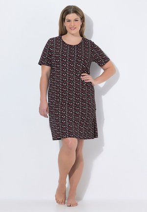 Kvinde står barfodet iført en sort knælængde kjole med korte ærmer og røde og hvide polkaprikker, smilende med en hånd på hoften.