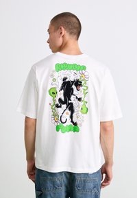Person som har på seg en hvit t-skjorte med et fargerikt bakdesign av en svart panter, hvite katter, grønne aliens, blomster og teksten "RIPNDIP PUMA".