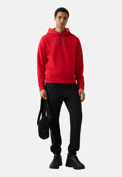 Jeune homme debout en sweat à capuche rouge, joggeurs noirs, baskets noires, tenant un petit sac noir avec une sangle devant un fond clair uni.